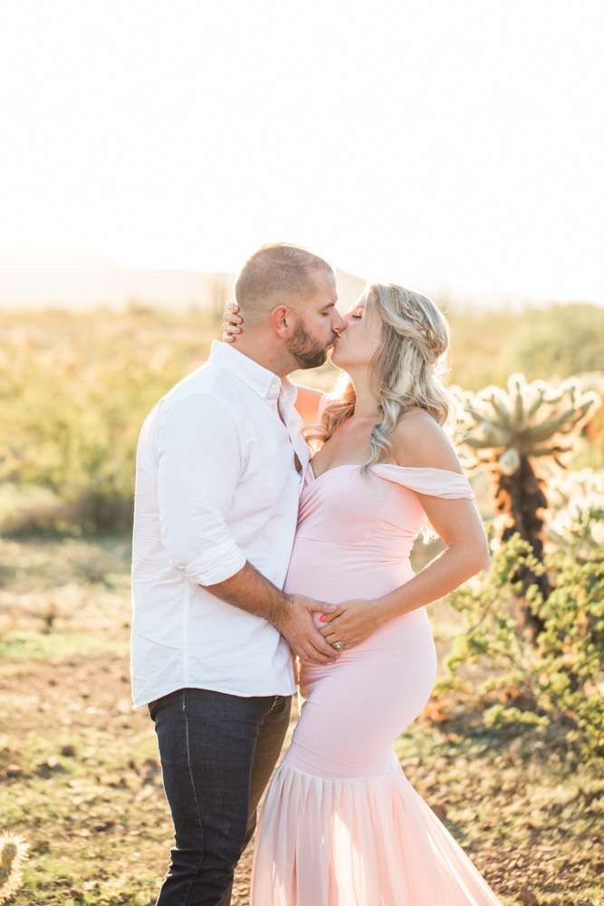 A couple kisses in front of a cactus during their maternity session with a Phoenix photographer.