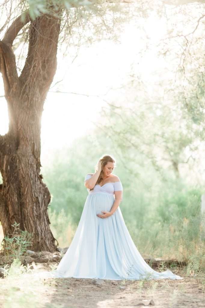 A North Phoenix maternity model poses in a blue dress in front of a tree.