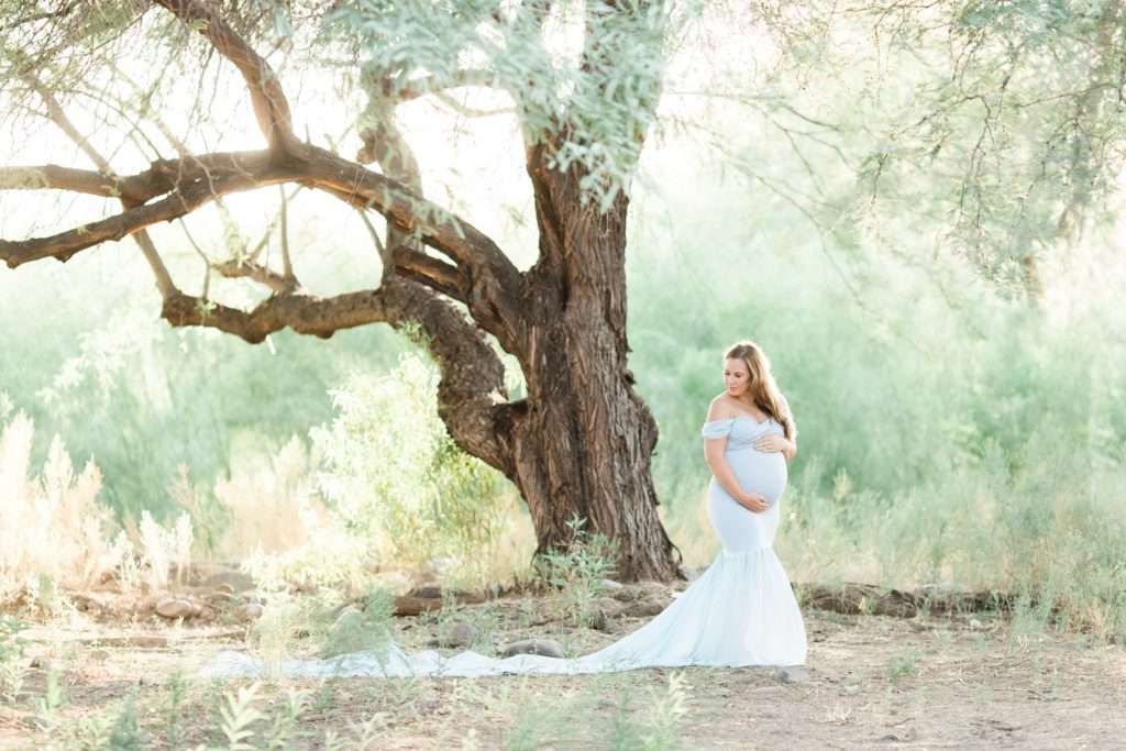A North Phoenix pregnant woman in a white dress is standing under a tree for her maternity photoshoot.