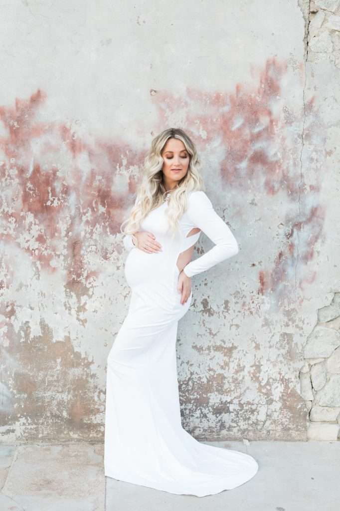 A pregnant woman in a white dress posing in front of a wall, captured by a Phoenix photographer.
