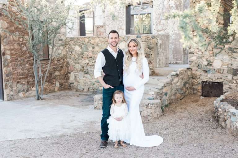A man and woman pose for a photo in front of a stone building captured by a maternity photographer in Phoenix.