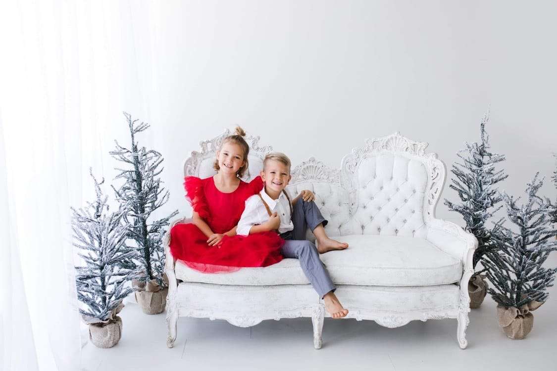 Newborn and maternity photographer capturing two children sitting on a white couch with Christmas trees in Phoenix.