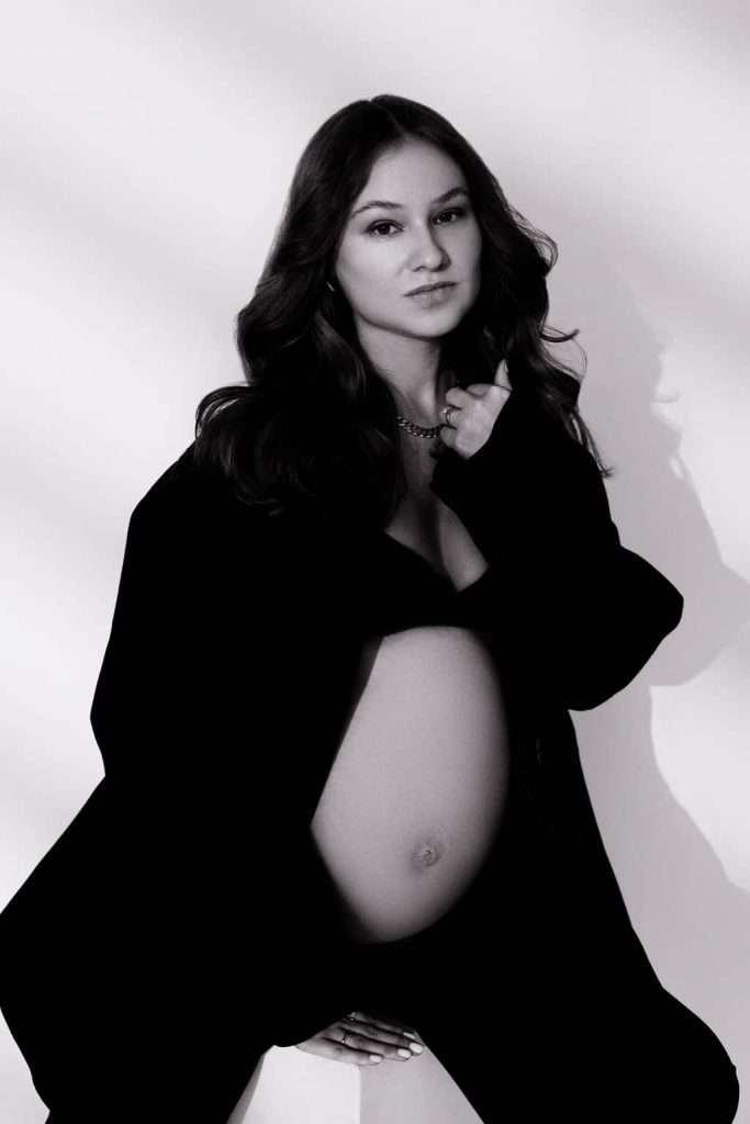 A pregnant woman is posing in front of a white wall.