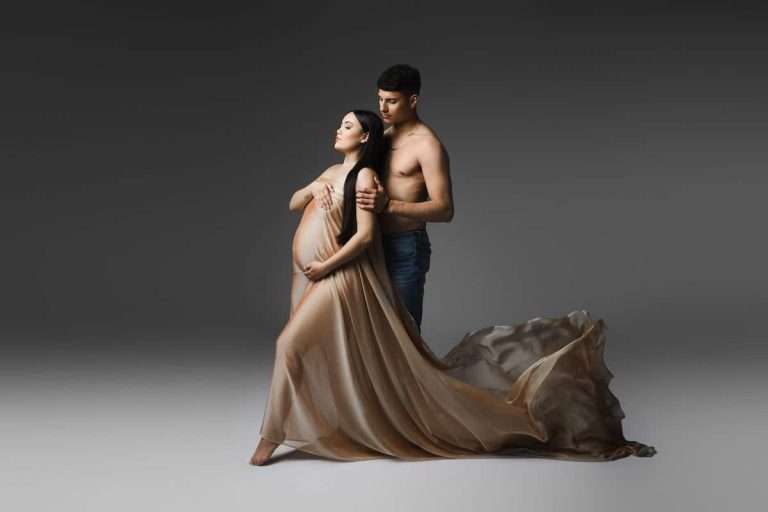 studio maternity A pregnant woman draped in flowing beige fabric stands with a bare-chested man embracing her, against a grey background.
