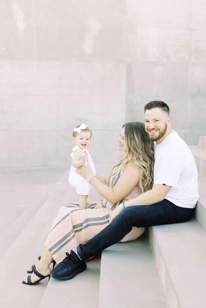 A couple sits on steps with a toddler. The woman holds the smiling child, who is standing in her lap. The man sits next to them, smiling at the camera.
