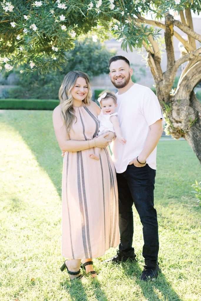 A woman in a light dress, a man in a white shirt and black pants, and a baby wearing a white outfit with a headband stand together on grass in front of a tree.