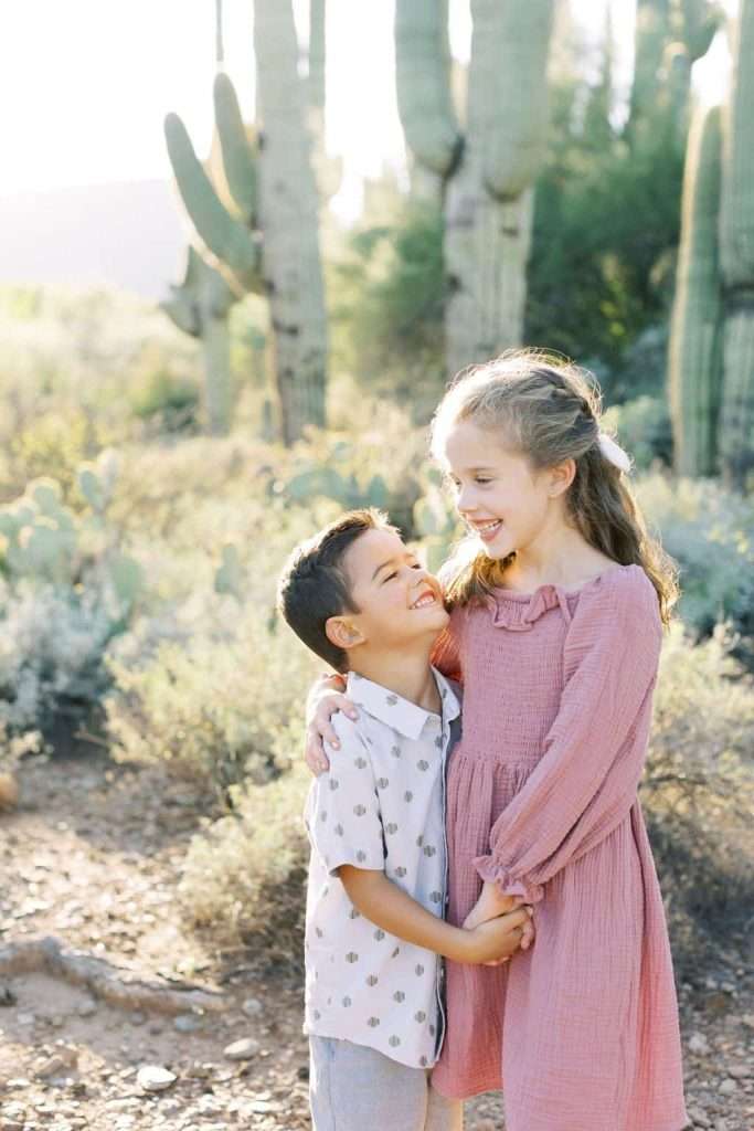 A young boy and girl stand smiling and holding hands in a desert landscape with tall cacti in the background. The boy wears a white shirt with a pattern, and the girl wears a pink dress.