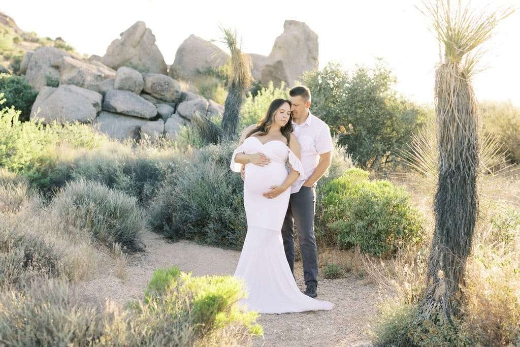 A couple poses in an outdoor setting with desert vegetation and rocks. The woman, wearing a white dress, is visibly pregnant, and the man stands behind her, holding her belly.