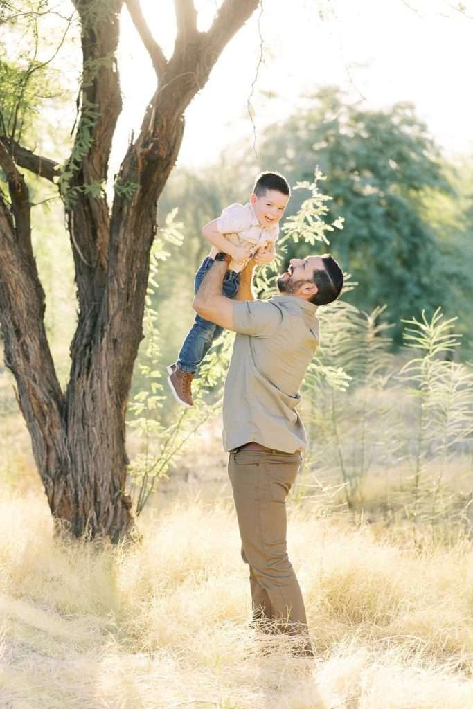 A bearded man in a beige shirt lifts a smiling young boy in a park with tall grass and trees in the background.