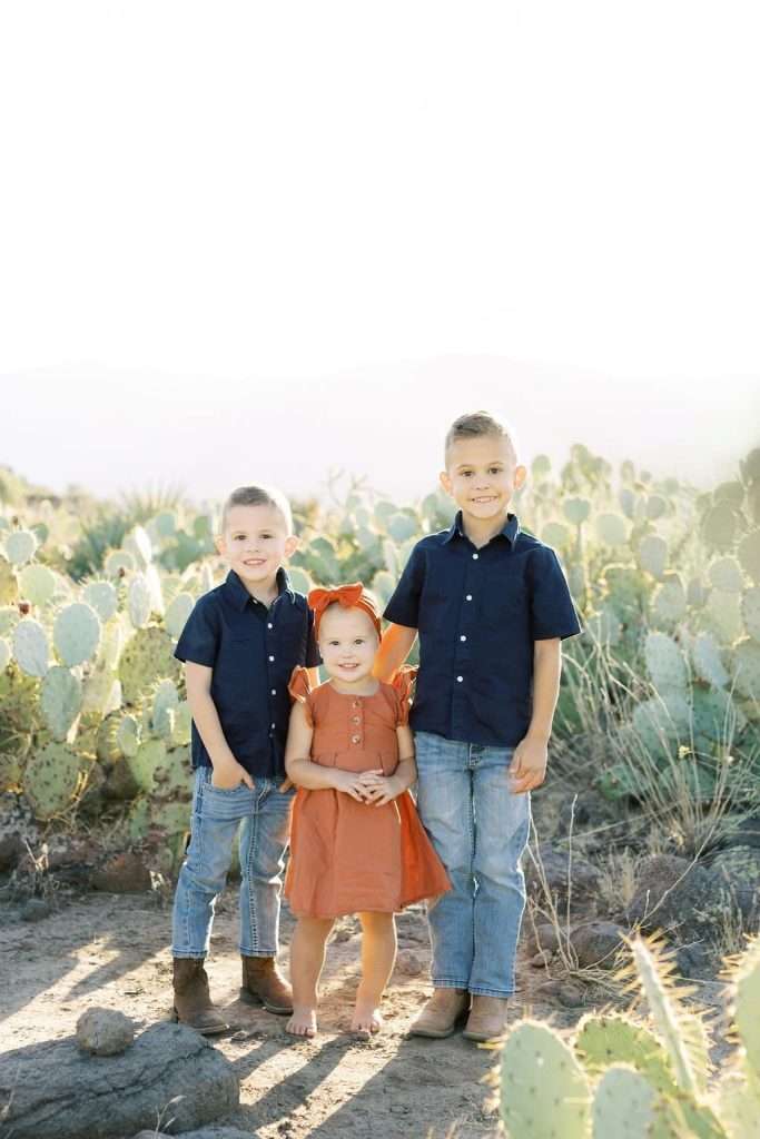 Three children are standing outside in a desert landscape with cacti. Two boys, wearing navy shirts and jeans, flank a younger girl in an orange dress. They are all smiling and looking at the camera.
