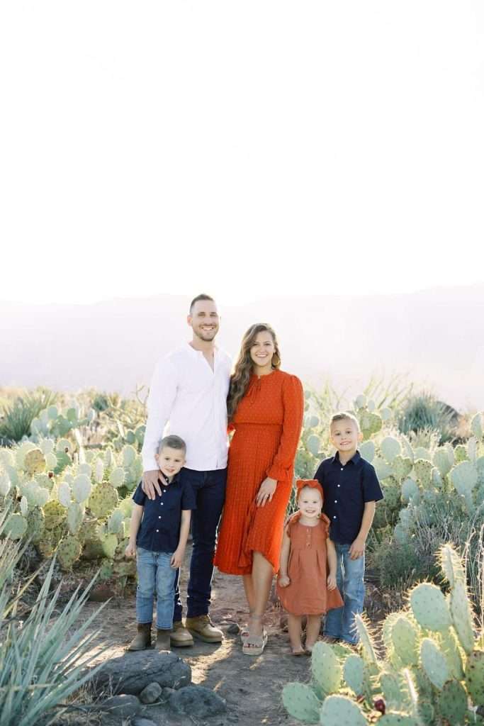 A family of five stands outside in a desert landscape with cacti. The man wears a white shirt, the woman a red dress, and the three children are in blue and orange outfits.