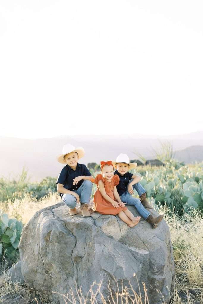 Three children wearing cowboy hats sit on a large rock in a desert landscape, with cacti and mountains in the background.