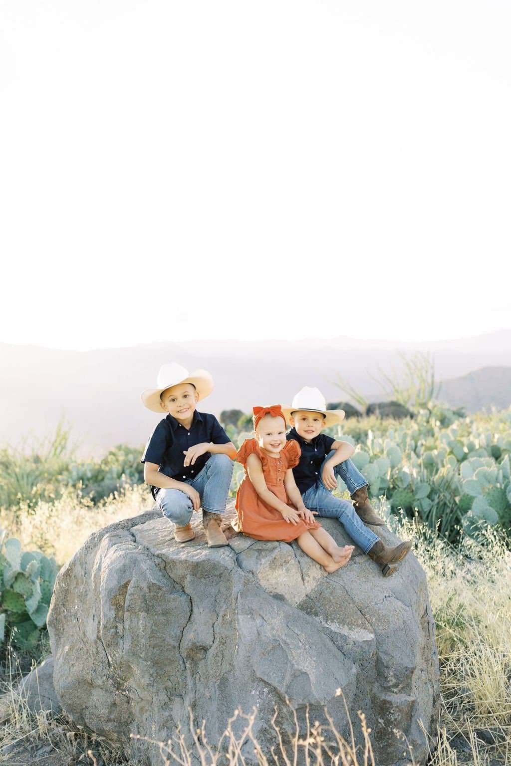 Three children wearing cowboy hats sit on a large rock in a desert landscape, with cacti and mountains in the background.