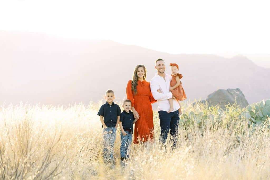 A family of five stands in a sunlit field with mountains in the background. The parents are dressed in white and red, while the three children wear dark tops and light pants.