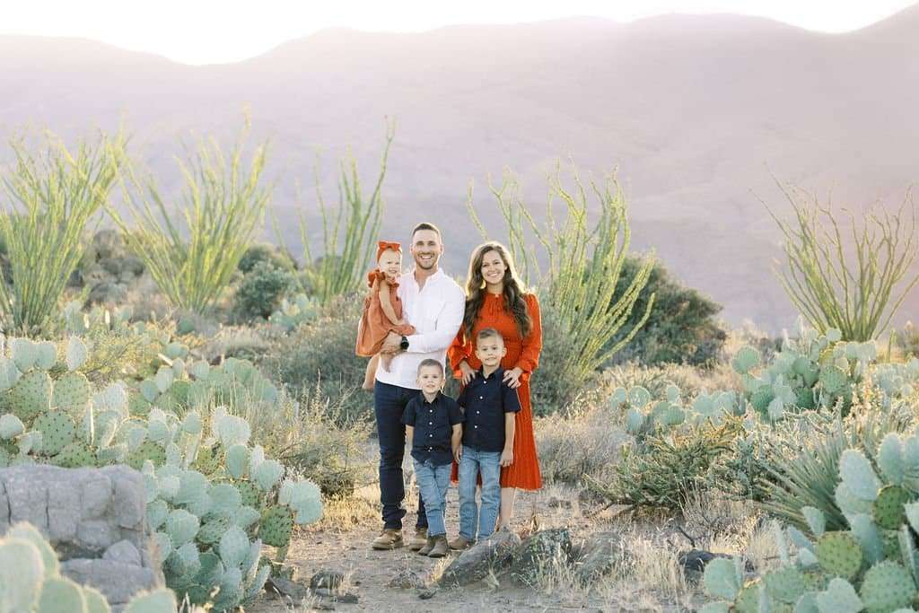 A family of five, including two adults and three young children, stand together in a desert landscape with cacti and mountains in the background.