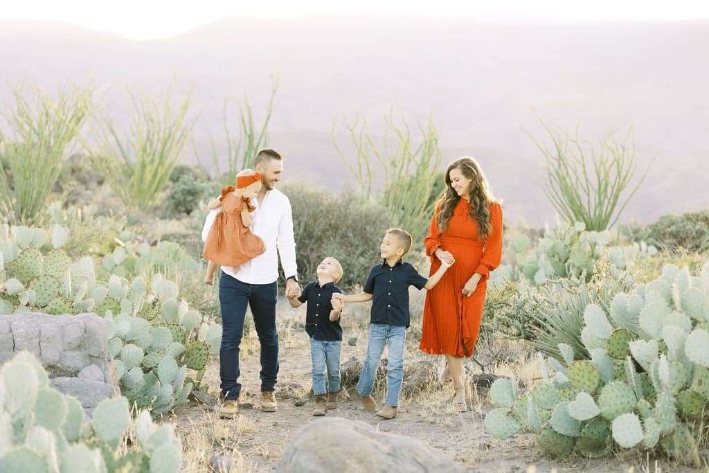 A family of five, dressed in autumn attire, walks together through a desert landscape with cactus plants, smiling and holding hands.