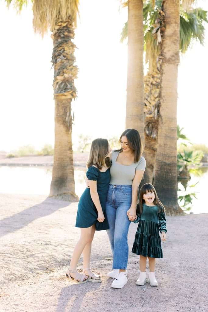 Three people stand together outdoors in front of palm trees; two are adults, and one is a child. One adult looks at the other, who is smiling at the camera while holding the child's hand.