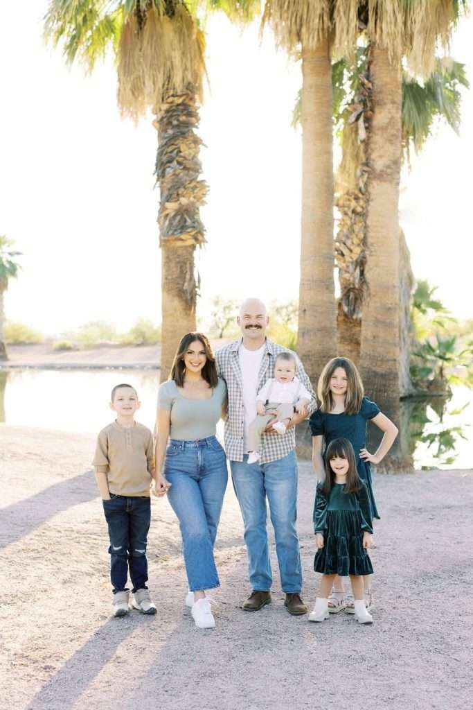 A family of six, including two adults and four children, poses outdoors in front of palm trees near a pond. The adults hold one baby and one child's hand, while the other two children stand nearby.