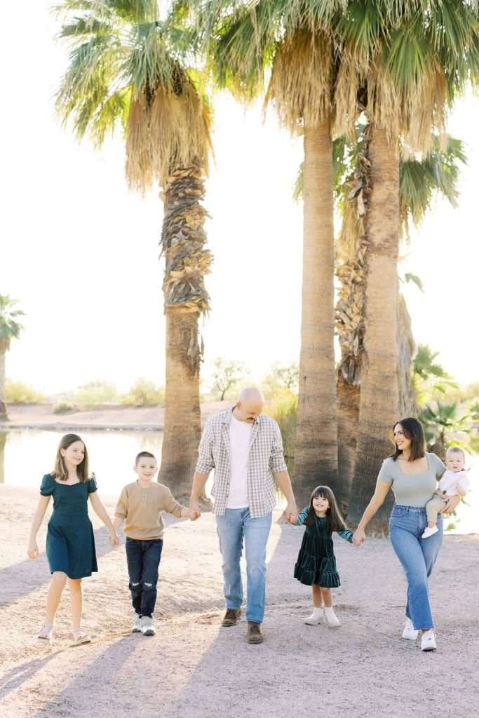 A family of five, including three children and two adults, walks hand in hand outdoors on a sunny day with tall palm trees in the background.