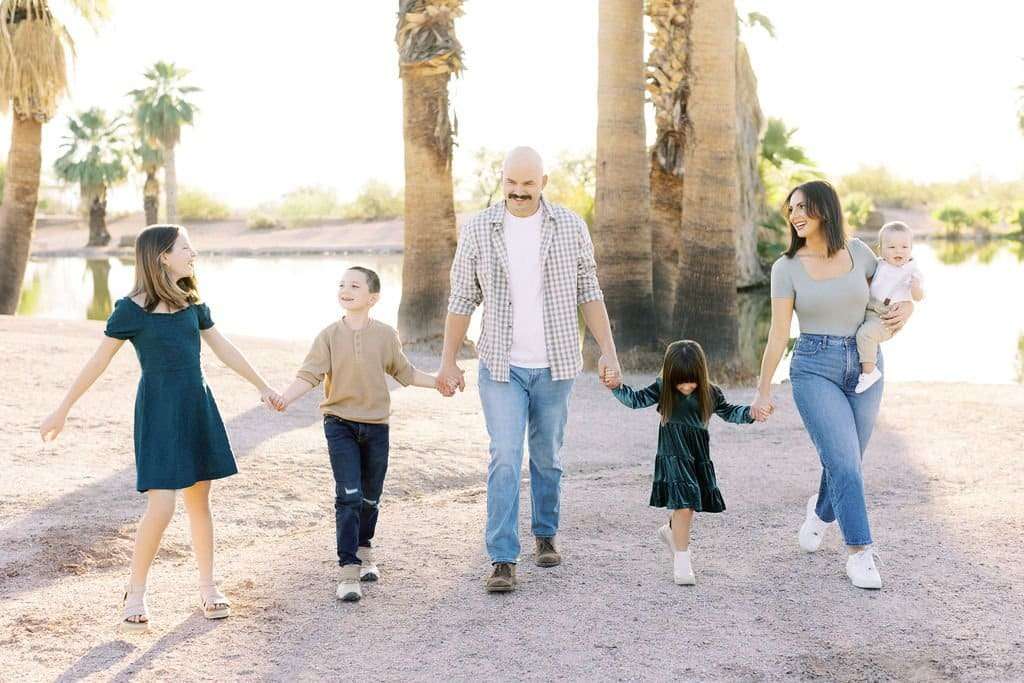 A family of six holding hands and walking outdoors. The group includes two adults and four children with a lake and palm trees in the background. One adult is carrying a baby.