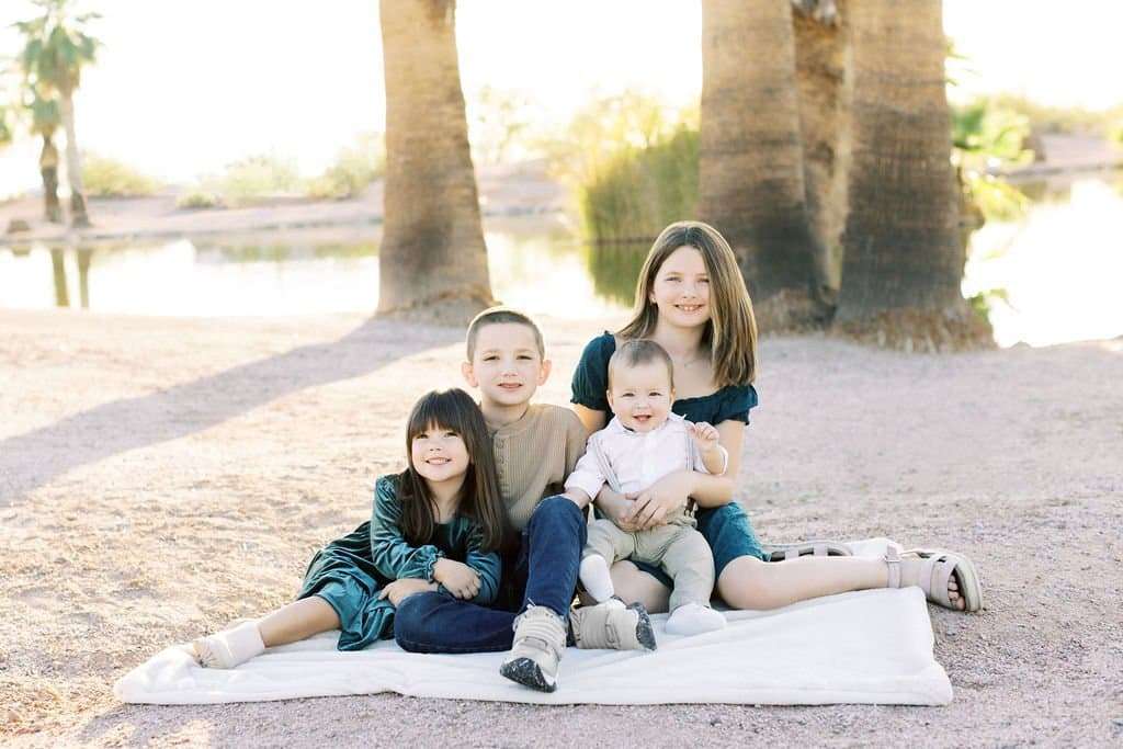 Four children sit on a white blanket outdoors, with three girls and one boy, smiling in front of palm trees and a pond.