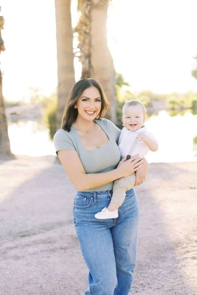 A woman in jeans and a light green top holds a smiling baby outdoors, with palm trees and a body of water in the background.