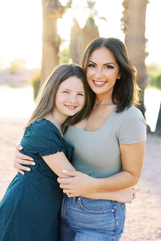 A woman and a young girl smile and pose for a photo outdoors with palm trees and sunlight in the background. The woman is wearing a light green top and jeans, while the girl is in a dark blue dress.