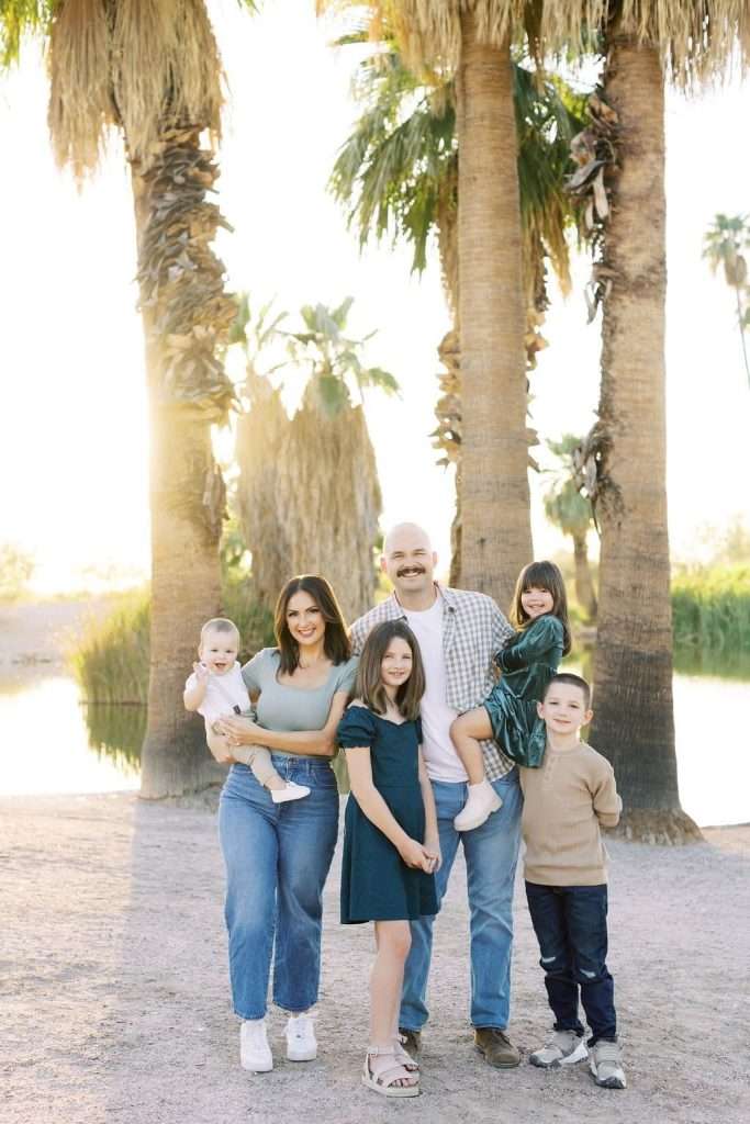 A family of six, including two adults and four children, poses outdoors in front of tall palm trees with a lake visible in the background. The adults stand behind the children, smiling at the camera.