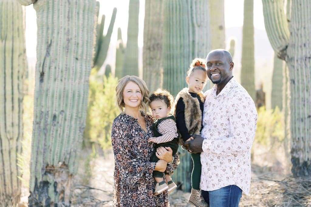 A family of four stands in front of tall cacti in a desert setting. The father holds a young child, and the mother holds a baby. Everyone is smiling.