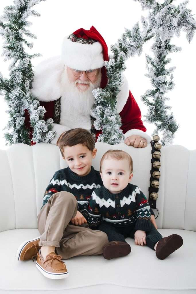 A person dressed as Santa Claus looks over a white sofa where two young children in festive sweaters sit. The scene is decorated with snowy garlands.