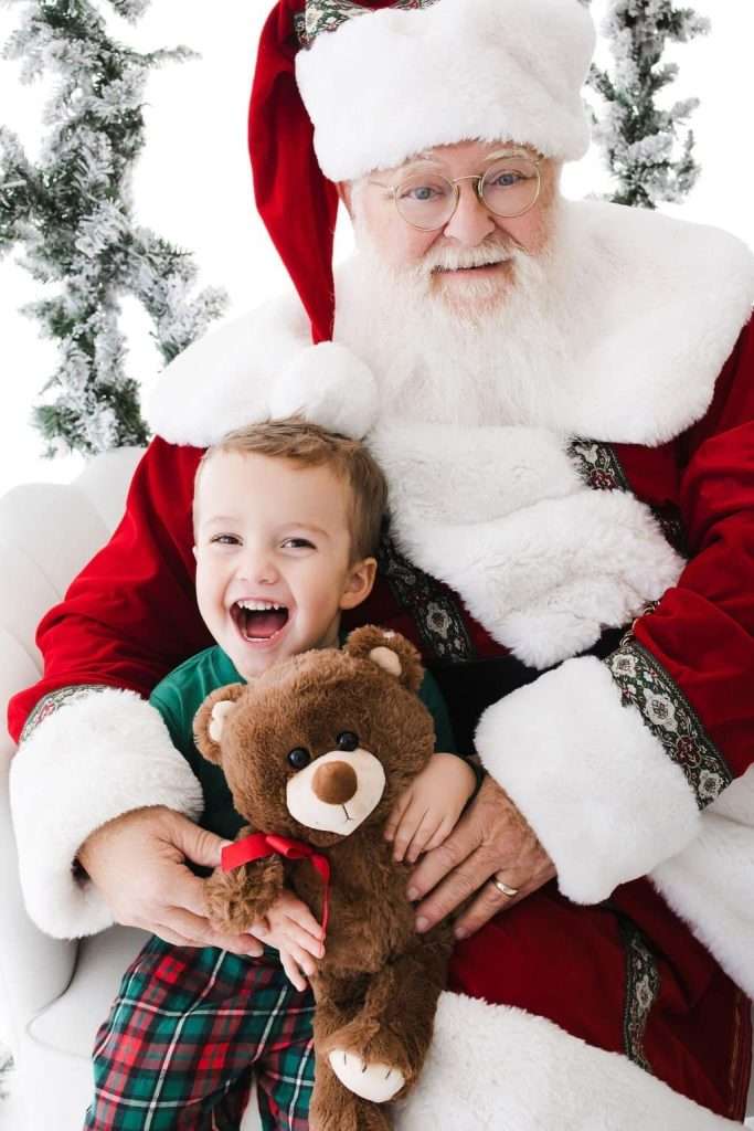 A smiling child holding a teddy bear sits on the lap of a person dressed as Santa Claus, with a festive background featuring decorated trees.