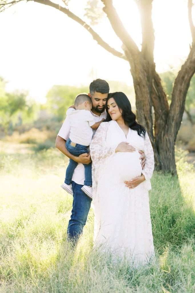 A pregnant woman in a white dress stands in a grassy field embraced by a man holding a young child. Sunlight filters through the trees in the background.