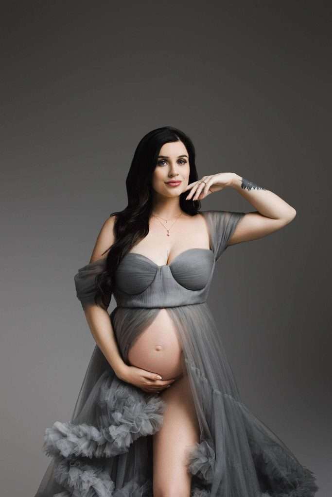maternity photographer phoenix - Pregnant woman in a flowing gray gown poses against a gray background, with one hand on her belly and the other raised to her face.