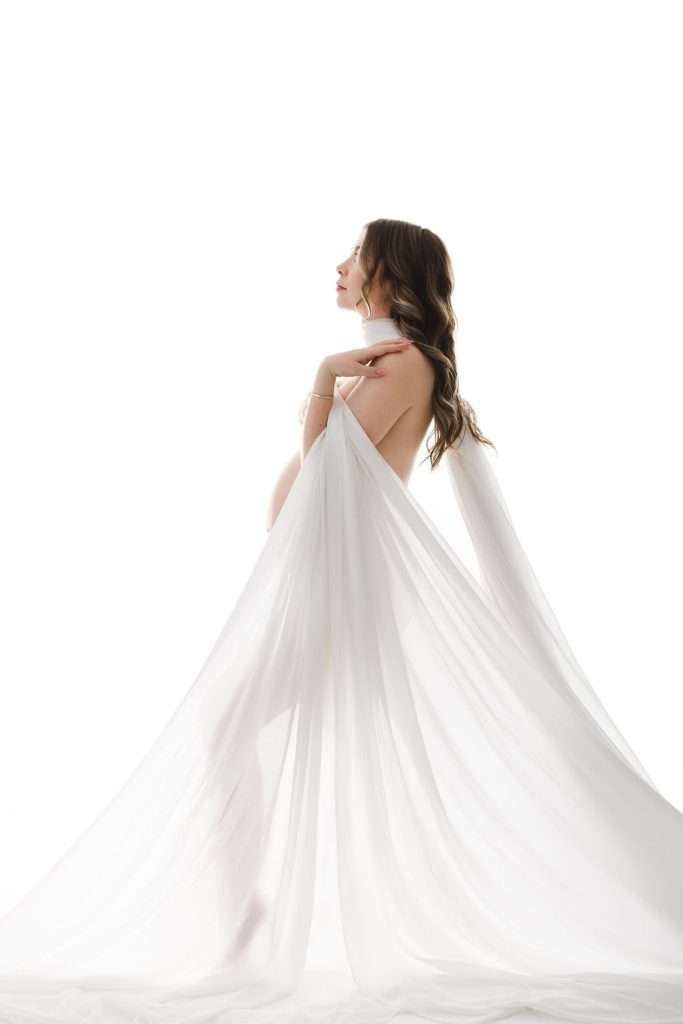 Pregnant woman standing in profile, draped in sheer white fabric, against a bright white background.