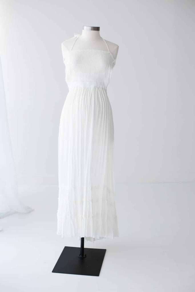 A white, sleeveless, ankle-length dress with a smocked bodice displayed on a dress form against a plain white background.
