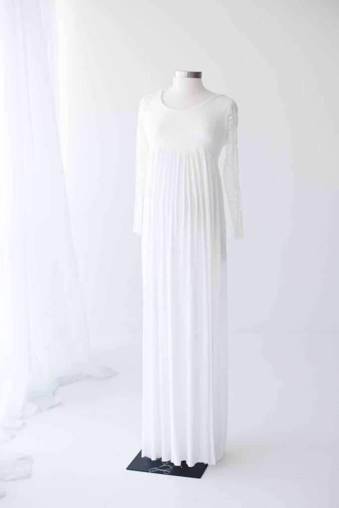 A long, white, pleated dress with long sleeves displayed on a dress form in a bright, minimalist room.