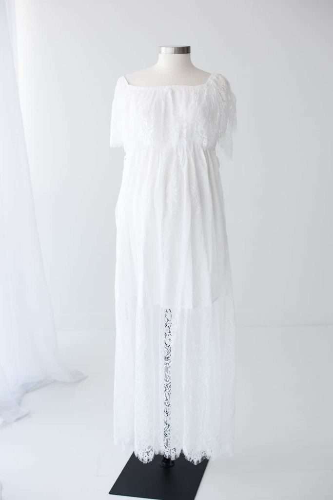 A white, off-the-shoulder, lace maternity dress is displayed on a dress form against a plain white background.