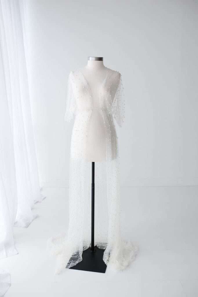 A sheer white dress with short sleeves and a long train, decorated with small beads, displayed on a dress form in a minimalist, white room.