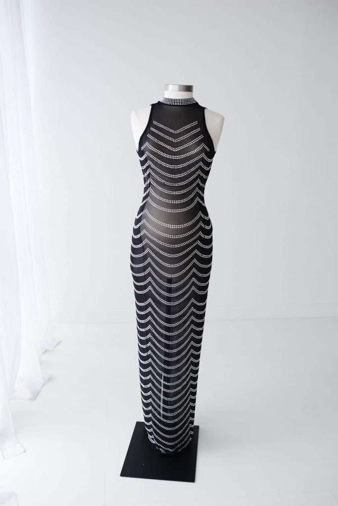 A sleeveless, floor-length black mesh dress with horizontal zigzag white stripe patterns displayed on a mannequin in a minimalist white room.