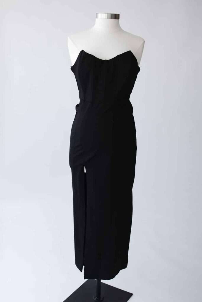Black strapless midi dress with fitted bodice and front slit, displayed on a white dress form against a plain background.