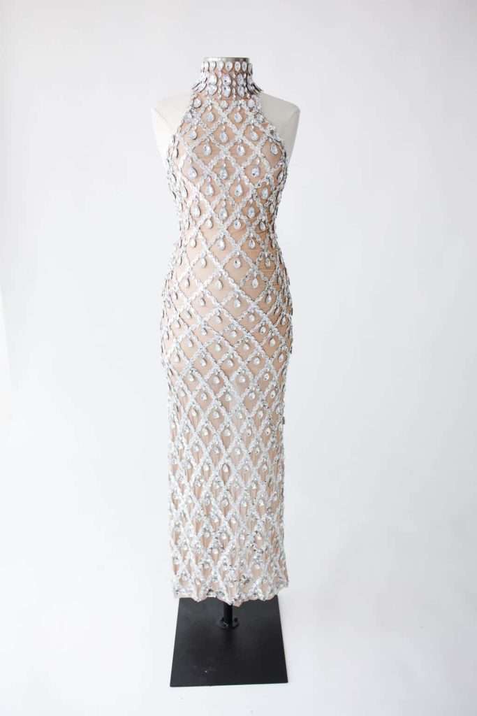 Sleeveless, floor-length nude dress with a high neckline, featuring a grid pattern of silver jeweled embellishments on a sheer fabric, displayed on a mannequin.