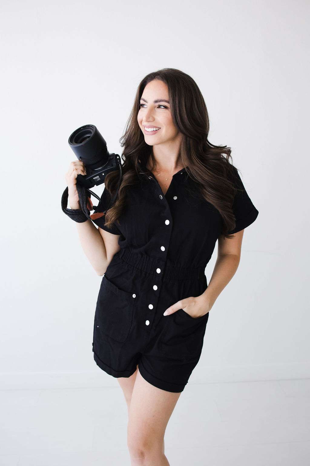 Amanda Capello, of Acapello Photography, stands against a white background with long brown hair, wearing a black button-up romper and holding a professional camera, smiling warmly.