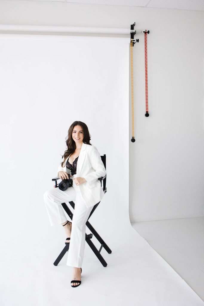 Woman in a white suit sits on a director's chair, holding a camera, in a bright photography studio—perfect for a maternity photographer Phoenix seeking creative backdrops with both white and colored options on the wall.
