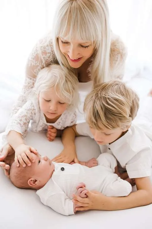 A woman with two young children gently touches and looks at a baby lying on a white surface. All are dressed in light-colored clothing, creating a serene moment captured by a Family Photographer Phoenix.