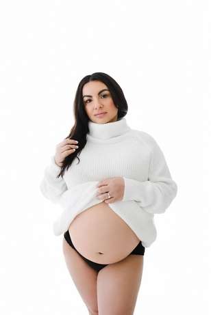 A pregnant woman in a white turtleneck sweater and black underwear stands against a white background, lifting her sweater to reveal her bare belly—captured beautifully by a Family Photographer Phoenix.