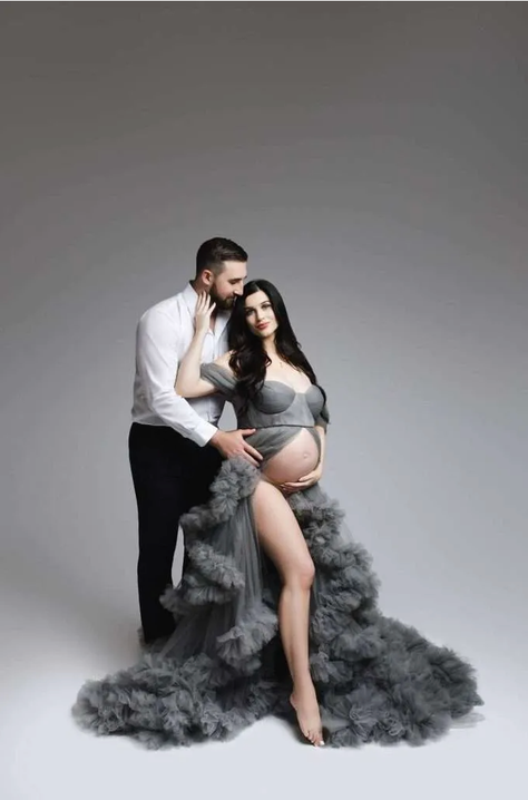 A man stands behind a pregnant woman wearing a sheer gray gown, with his hands on her belly, posing together in a studio setting—captured beautifully by a Family Photographer Phoenix.