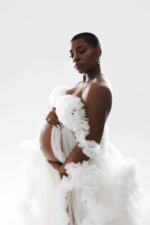 A pregnant woman in a white, floral, sheer gown poses holding her belly against a plain white background in this Acapello Photography portrait by Amanda Capello.