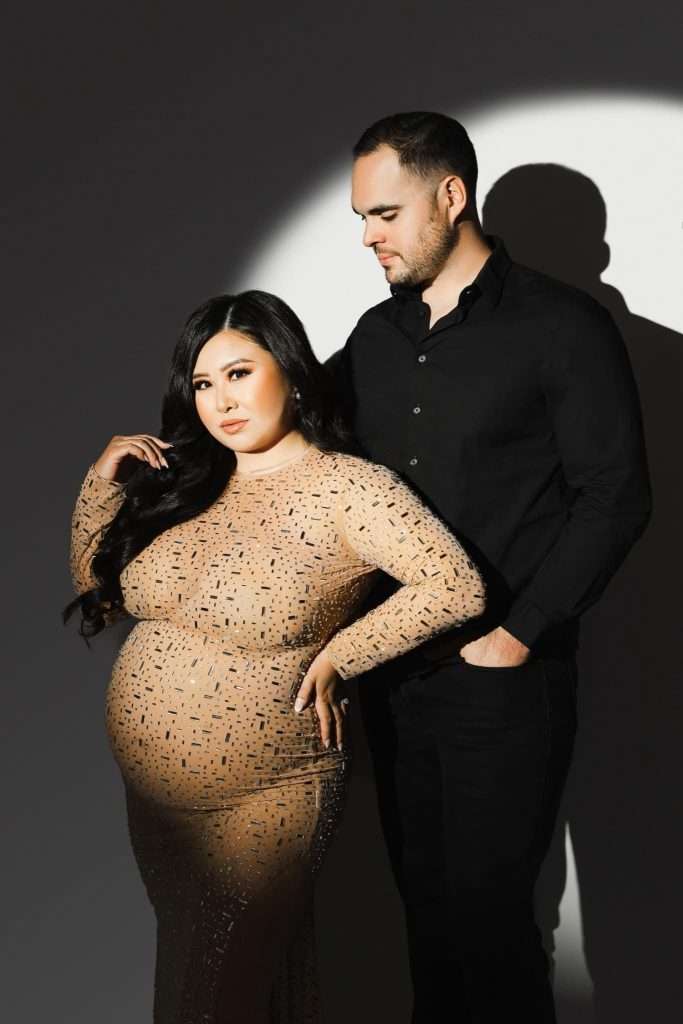A pregnant woman in a beige, sequined dress stands beside a man in black attire, both posing under a spotlight—an elegant moment captured by a skilled maternity photographer Phoenix couples trust.