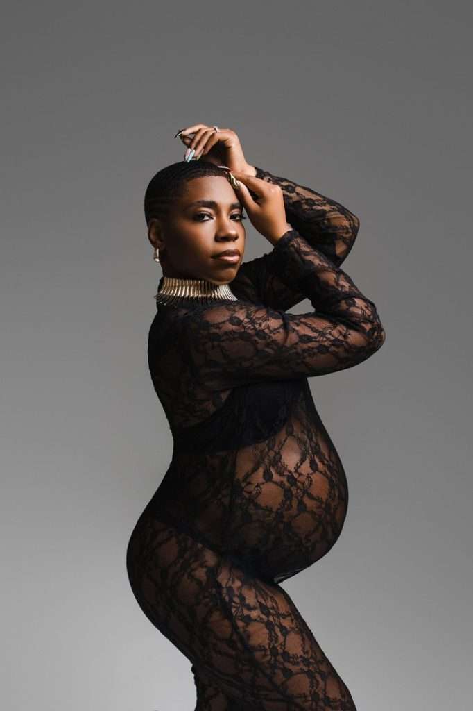 A pregnant woman poses confidently in a black lace bodysuit against a plain gray background, wearing a statement necklace and holding her hands above her head—captured beautifully by a talented maternity photographer Phoenix.