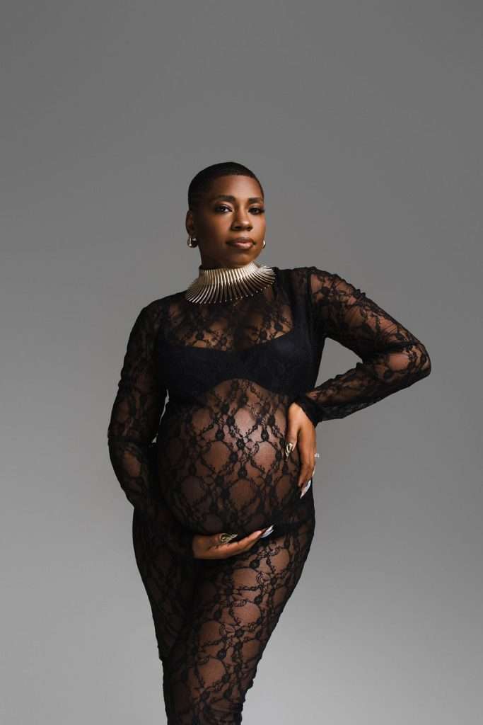 A pregnant woman in a sheer black lace outfit poses with one hand on her hip and the other supporting her belly, captured by a Family Photographer Phoenix, against a plain gray background.